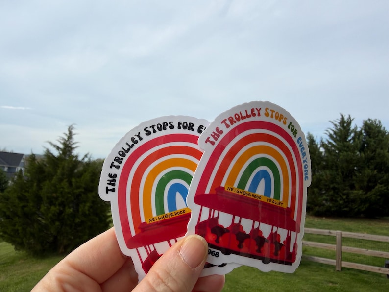 Pu&ograve; includere: Due adesivi con un design arcobaleno e la scritta "The Trolley Stops for Everyone". Gli adesivi presentano un tram rosso e un arco arcobaleno. &Egrave; visibile anche la parola "Neighborhood".