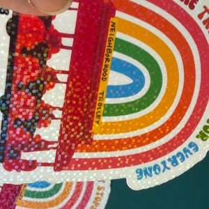 May include: A sticker with a rainbow design and a trolley illustration. The text on the sticker says "The Trolley Stops for Everyone" and "Neighborhood Trolley". The sticker has a glittery, iridescent finish.