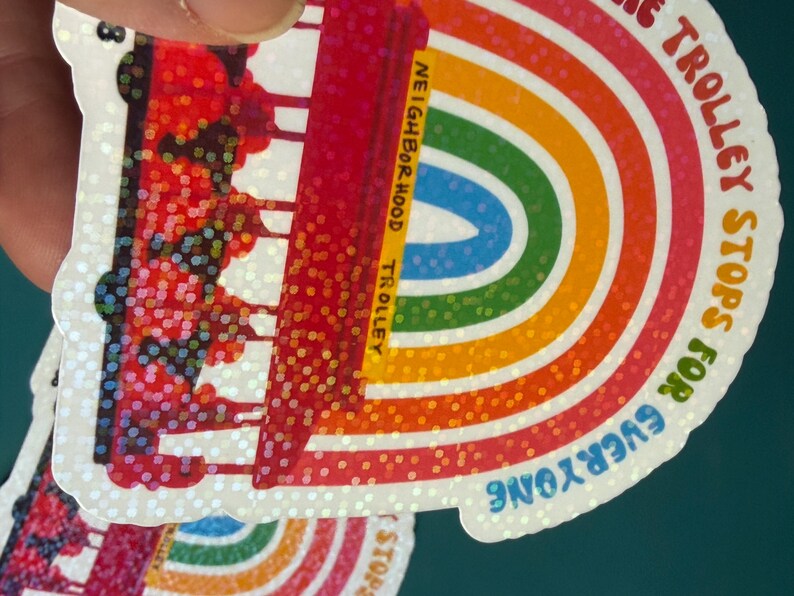 May include: A sticker with a rainbow design and a trolley illustration. The text on the sticker says "The Trolley Stops for Everyone" and "Neighborhood Trolley". The sticker has a glittery, iridescent finish.
