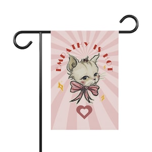 May include: A garden flag featuring a vintage-style illustration of a cat with a pink bow, set against a pink and white striped background. The text "EMPATHY IS HOT" is arched above the cat, with a heart and stars below.