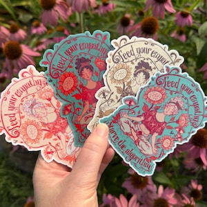 May include: Three die-cut stickers featuring a person in a meditative pose surrounded by sunflowers and floral motifs. The text reads Feed your empathy and Starve the algorithm. The stickers are rendered in color palettes of teal, red, and cream.