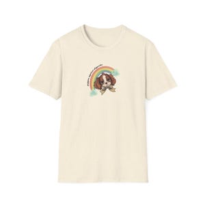Puede incluir: Camiseta color crema con un gráfico de un Cavalier King Charles Spaniel, un arcoíris y el texto "puppies against oligarchs". El perro tiene pelaje marrón y blanco y lleva una pajarita.