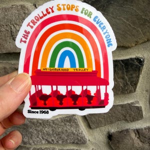 Pu&ograve; includere: Un adesivo colorato con un tram rosso con passeggeri, ambientato su un arcobaleno. Il testo "THE TROLLEY STOPS FOR EVERYONE" &egrave; sopra l'arcobaleno, con "NEIGHBORHOOD TROLLEY" e "Since 1968" sotto. L'adesivo &egrave; tenuto contro un muro di pietra.