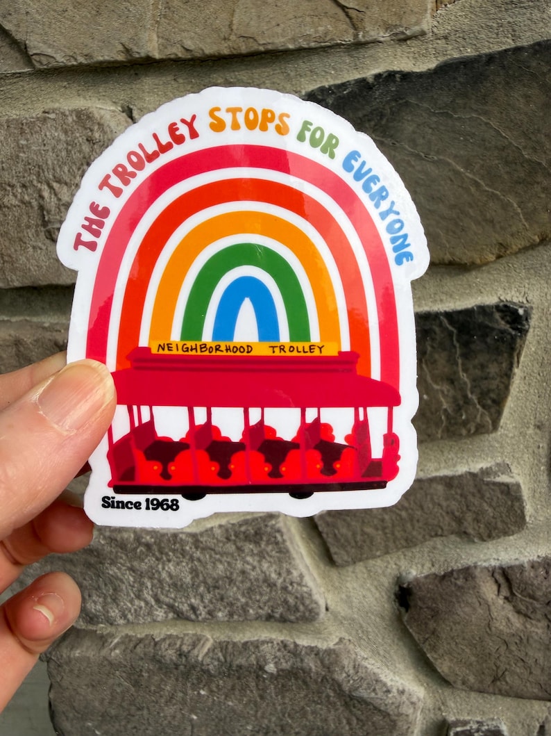 Pu&ograve; includere: Un adesivo colorato con un tram rosso con passeggeri, ambientato su un arcobaleno. Il testo "THE TROLLEY STOPS FOR EVERYONE" &egrave; sopra l'arcobaleno, con "NEIGHBORHOOD TROLLEY" e "Since 1968" sotto. L'adesivo &egrave; tenuto contro un muro di pietra.