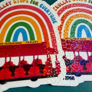 May include: Two stickers featuring a rainbow design and a red trolley. The stickers display the text "Trolley Stops for Everyone" and "Neighborhood Trolley". One sticker has the year "1968". The stickers have a glittery finish.