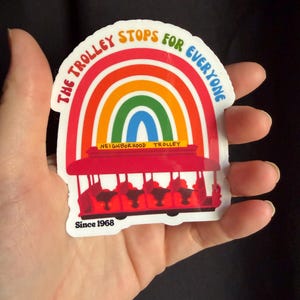 Pu&ograve; includere: Un adesivo colorato raffigurante un tram rosso e un arcobaleno, con la scritta "THE TROLLEY STOPS FOR EVERYONE". Include anche "NEIGHBORHOOD TROLLEY" e "Since 1968". L'adesivo &egrave; tenuto in mano.