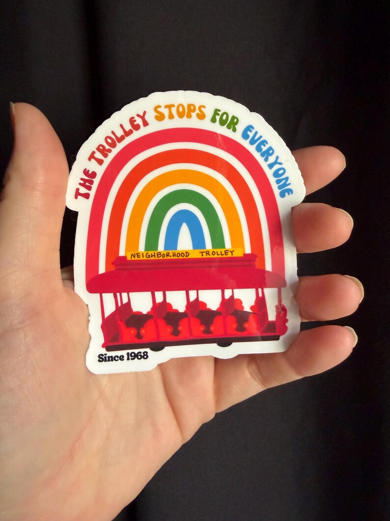 Pu&ograve; includere: Un adesivo colorato raffigurante un tram rosso e un arcobaleno, con la scritta "THE TROLLEY STOPS FOR EVERYONE". Include anche "NEIGHBORHOOD TROLLEY" e "Since 1968". L'adesivo &egrave; tenuto in mano.