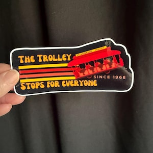 Puede incluir: Una pegatina con una ilustraci&oacute;n de tranv&iacute;a rojo y el texto "THE TROLLEY STOPS FOR EVERYONE SINCE 1968". El dise&ntilde;o incluye rayas amarillas, naranjas y rojas sobre un fondo negro.