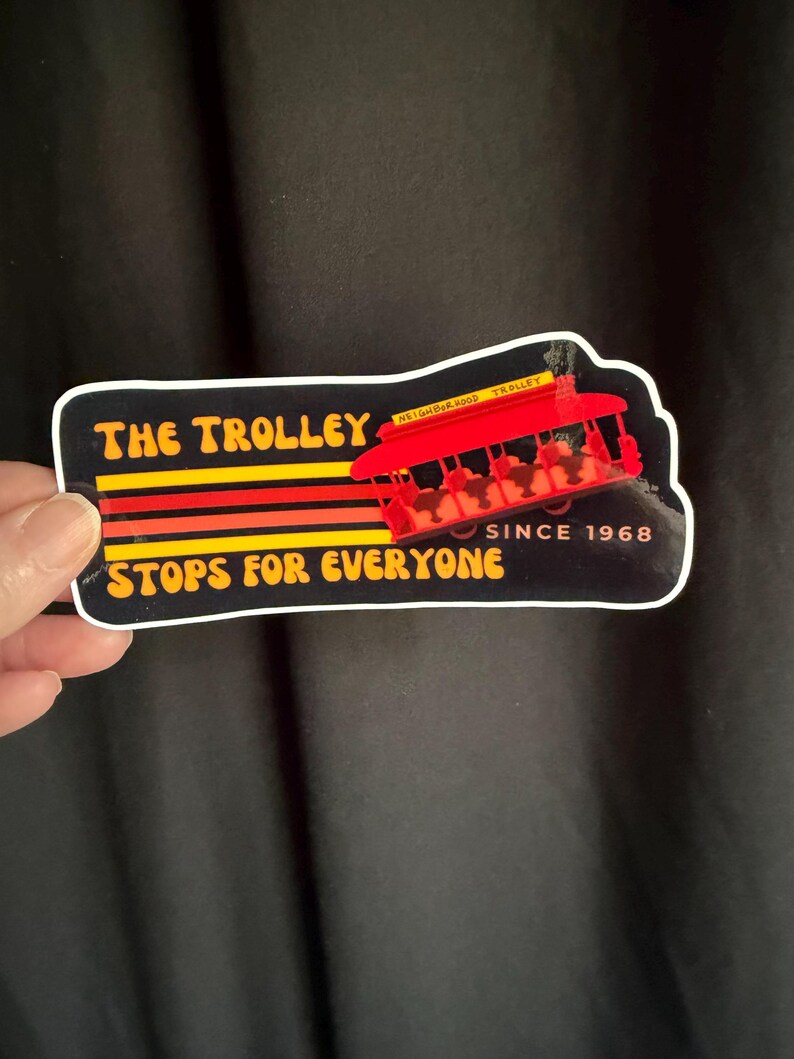 Puede incluir: Una pegatina con una ilustraci&oacute;n de tranv&iacute;a rojo y el texto "THE TROLLEY STOPS FOR EVERYONE SINCE 1968". El dise&ntilde;o incluye rayas amarillas, naranjas y rojas sobre un fondo negro.