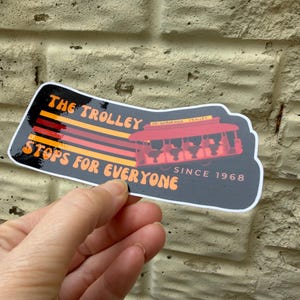 Puede incluir: Una pegatina con el texto "THE TROLLEY STOPS FOR EVERYONE SINCE 1968." La pegatina presenta un gr&aacute;fico de tranv&iacute;a rojo con pasajeros. El texto es naranja y amarillo sobre un fondo negro.