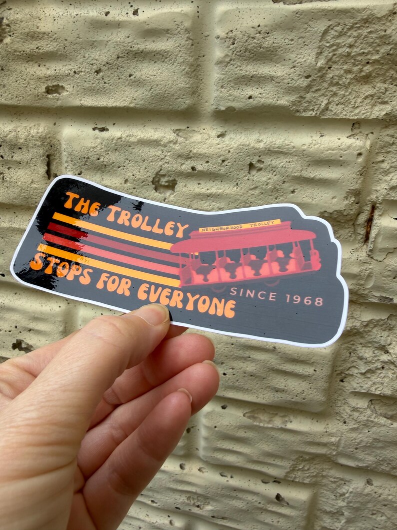 Puede incluir: Una pegatina con el texto "THE TROLLEY STOPS FOR EVERYONE SINCE 1968." La pegatina presenta un gr&aacute;fico de tranv&iacute;a rojo con pasajeros. El texto es naranja y amarillo sobre un fondo negro.