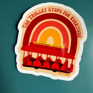 May include: A sticker featuring a red trolley with a rainbow design in shades of red, orange, and yellow. The text "THE TROLLEY STOPS FOR EVERYONE" arches above the trolley. The words "NEIGHBORHOOD TROLLEY" are on the trolley, and "Since 1968" is at the bottom.