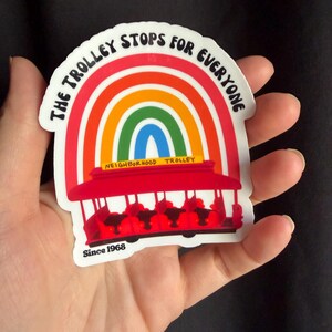 Pu&ograve; includere: Un adesivo raffigurante un tram rosso con un arcobaleno sopra. Il testo "THE TROLLEY STOPS FOR EVERYONE" &egrave; arcuato sopra l'arcobaleno. L'adesivo include anche il testo "NEIGHBORHOOD TROLLEY" e "Since 1968".