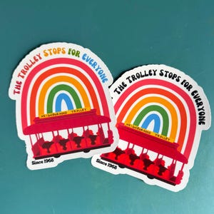 Pu&ograve; includere: Due adesivi con un design arcobaleno e un tram rosso. Il testo sugli adesivi recita "THE TROLLEY STOPS FOR EVERYONE" e "NEIGHBORHOOD TROLLEY" con "Since 1968" in basso.