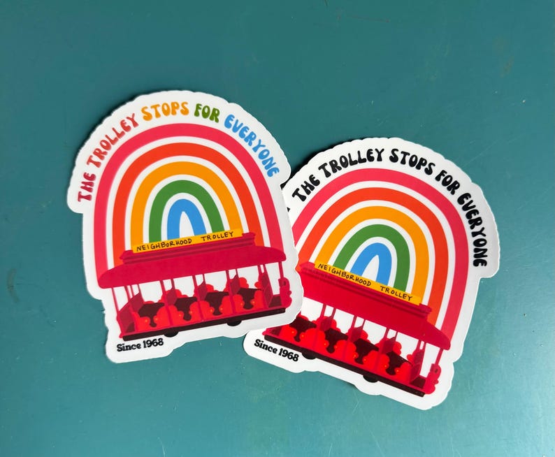 Pu&ograve; includere: Due adesivi con un design arcobaleno e un tram rosso. Il testo sugli adesivi recita "THE TROLLEY STOPS FOR EVERYONE" e "NEIGHBORHOOD TROLLEY" con "Since 1968" in basso.