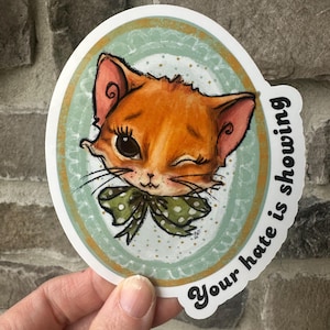 Könnte beinhalten: Ein Aufkleber mit einer Illustration einer zwinkernden, orangefarbenen Katze mit einer grünen Schleife mit weißen Punkten. Die Katze befindet sich vor einem hellgrünen und goldenen ovalen Hintergrund. Der Text "Your hate is showing" ist aufgedruckt.