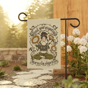 May include: A rectangular garden flag featuring a cream background with a central illustration of a person sitting in a lotus position surrounded by sunflowers. The text reads Feed your empathy and Starve the algorithm in dark brown script.