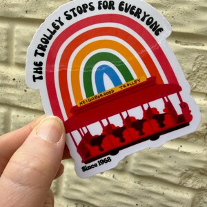 Pu&ograve; includere: Un adesivo con un tram rosso e un arcobaleno. Il testo "THE TROLLEY STOPS FOR EVERYONE" &egrave; in alto, con "NEIGHBORHOOD TROLLEY" e "Since 1968" in basso. L'adesivo &egrave; tenuto in mano.