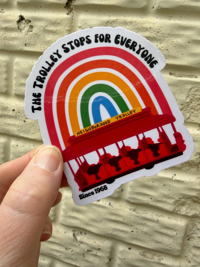 Pu&ograve; includere: Un adesivo con un tram rosso e un arcobaleno. Il testo "THE TROLLEY STOPS FOR EVERYONE" &egrave; in alto, con "NEIGHBORHOOD TROLLEY" e "Since 1968" in basso. L'adesivo &egrave; tenuto in mano.