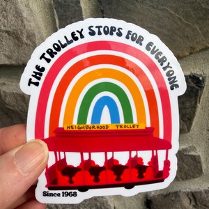 Pu&ograve; includere: Un adesivo con un design arcobaleno e un'illustrazione di tram rosso. Il testo recita "The Trolley Stops For Everyone" e "Neighborhood Trolley Since 1968". L'adesivo ha un bordo bianco ed &egrave; tenuto in mano.