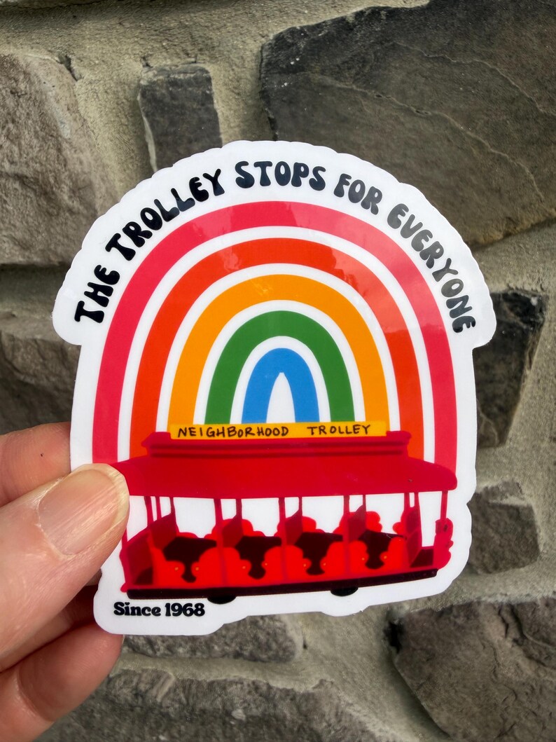 Pu&ograve; includere: Un adesivo con un design arcobaleno e un'illustrazione di tram rosso. Il testo recita "The Trolley Stops For Everyone" e "Neighborhood Trolley Since 1968". L'adesivo ha un bordo bianco ed &egrave; tenuto in mano.