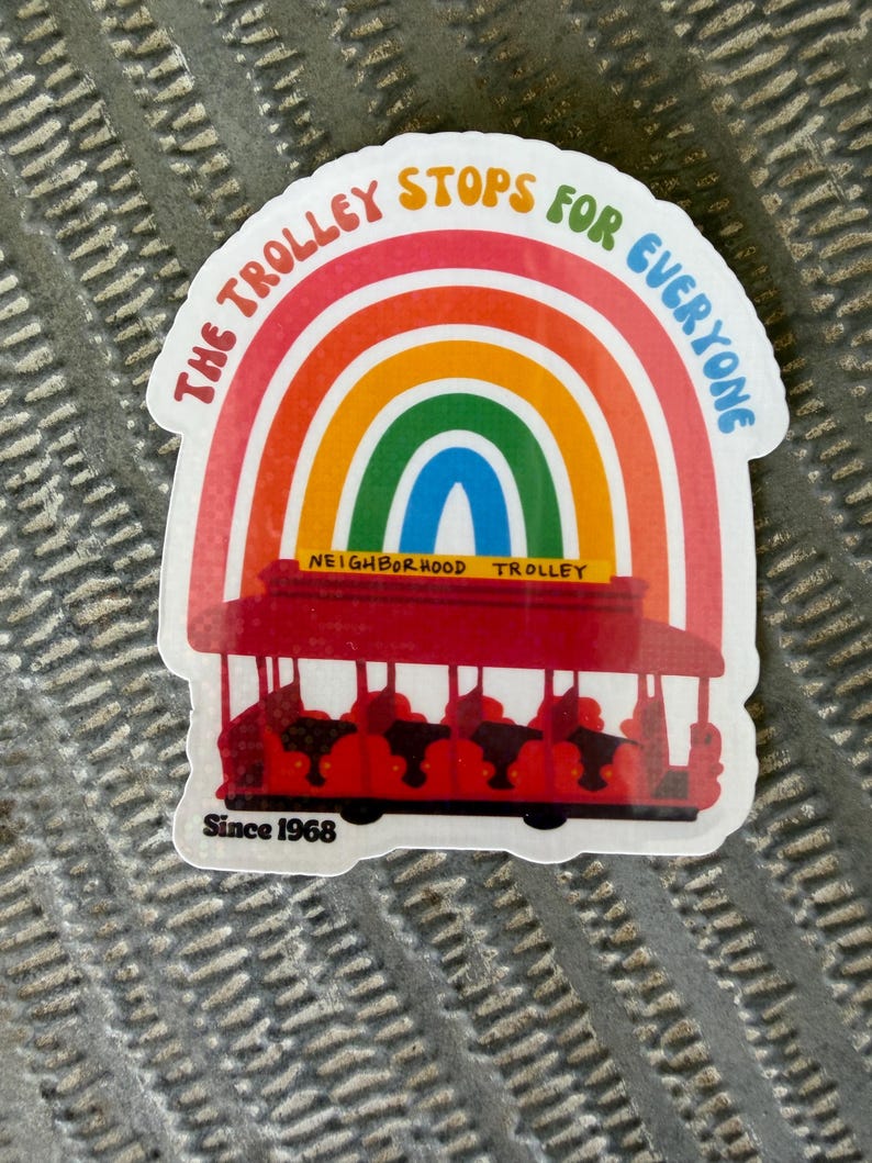 May include: A vibrant sticker with a red trolley design under a rainbow. The text "THE TROLLEY STOPS FOR EVERYONE" is above the rainbow. "NEIGHBORHOOD TROLLEY" is below, and "Since 1968" is at the bottom. A cheerful, colourful design.