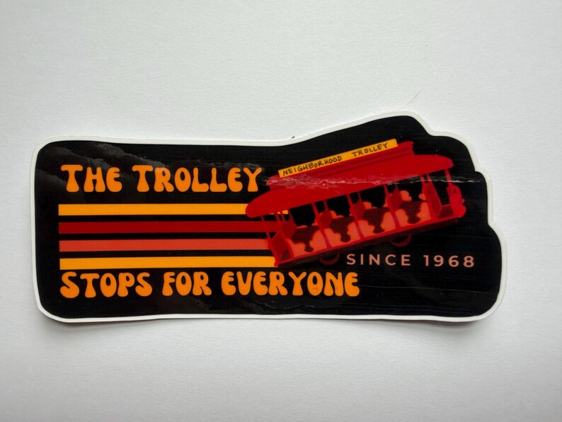 Puede incluir: Una pegatina con fondo negro y las palabras "The Trolley Stops For Everyone" en naranja y amarillo. Un gr&aacute;fico de tranv&iacute;a rojo est&aacute; a la derecha, con el texto "Neighborhood Trolley" encima. La pegatina dice "Since 1968".