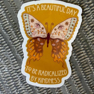 Op de afbeelding: Een gestanste sticker met een vlinder met ingewikkelde patronen, waaronder ogen en bloemmotieven in tinten oranje, crème en bruin. De tekst "IT'S A BEAUTIFUL DAY TO BE RADICALIZED BY KINDNESS" is in het wit rond de vlinder gedrukt.