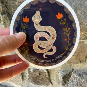 May include: A circular sticker featuring a light grey snake with red floral patterns on a dark blue background. Two orange flowers flank the snake. The text THE ALGORITHM IS POISON is printed in white along the bottom edge of the circular design.