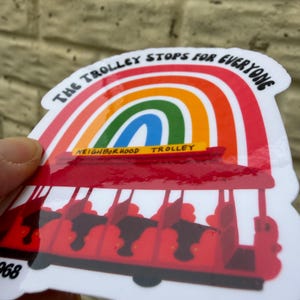Pu&ograve; includere: Un adesivo raffigurante un tram rosso sotto un arcobaleno, con la scritta "THE TROLLEY STOPS FOR EVERYONE". Il tram ha sedili neri ed &egrave; etichettato "NEIGHBORHOOD TROLLEY" in giallo. L'adesivo ha un bordo bianco.