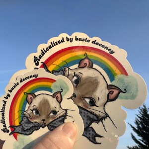 K&ouml;nnte beinhalten: Mehrere Aufkleber mit einer Cartoon-Katze, einem Regenbogen und dem Text "Radicalized by basic decency". Die Katze tr&auml;gt eine graue Fliege. Der Hintergrund ist ein klarer blauer Himmel.