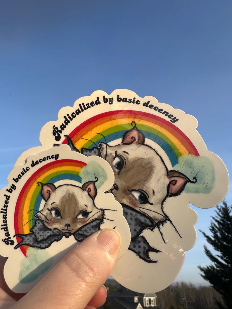 K&ouml;nnte beinhalten: Mehrere Aufkleber mit einer Cartoon-Katze, einem Regenbogen und dem Text "Radicalized by basic decency". Die Katze tr&auml;gt eine graue Fliege. Der Hintergrund ist ein klarer blauer Himmel.