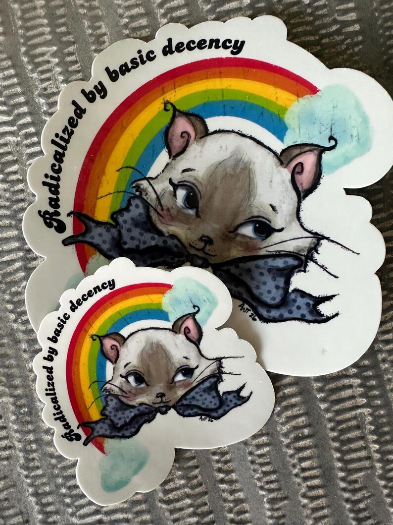 K&ouml;nnte beinhalten: Zwei Aufkleber mit einer Cartoon-Katze mit blauen Augen, einer Schleife und einem Regenbogen. Der Text "radicalized by basic decency" ist um die Katze und den Regenbogen herum angeordnet. Die Aufkleber sind wei&szlig; mit einem wolkenf&ouml;rmigen Rand.