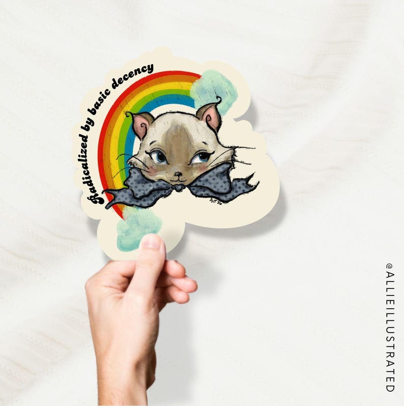 Waterproof vinyl sticker featuring Prudence the Siamese cat with blue eyes and grey polka dot bow, backed by a vibrant rainbow with soft clouds on a cream scalloped background. Arched text reads Radicalized by Basic Decency. Original hand-drawn art by Allison Weeks Thomas of The Rising Up Collective