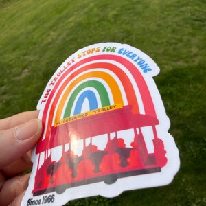 Pu&ograve; includere: Un adesivo colorato raffigurante un tram rosso con un arco a forma di arcobaleno. Il testo recita "The Trolley Stops for Everyone" e "Neighborhood Trolley". L'adesivo include anche "Since 1968".