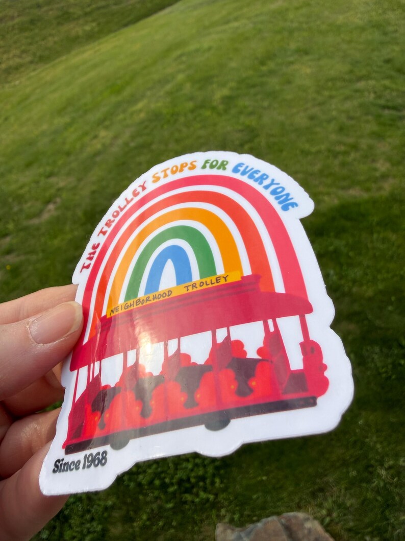 Pu&ograve; includere: Un adesivo colorato raffigurante un tram rosso con un arco a forma di arcobaleno. Il testo recita "The Trolley Stops for Everyone" e "Neighborhood Trolley". L'adesivo include anche "Since 1968".