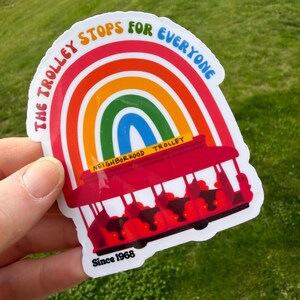 Pu&ograve; includere: Un adesivo colorato raffigurante un tram rosso con passeggeri, su uno sfondo arcobaleno. Il testo recita "THE TROLLEY STOPS FOR EVERYONE" e "NEIGHBORHOOD TROLLEY", con "Since 1968" in basso. L'adesivo &egrave; tenuto su uno sfondo erboso verde.