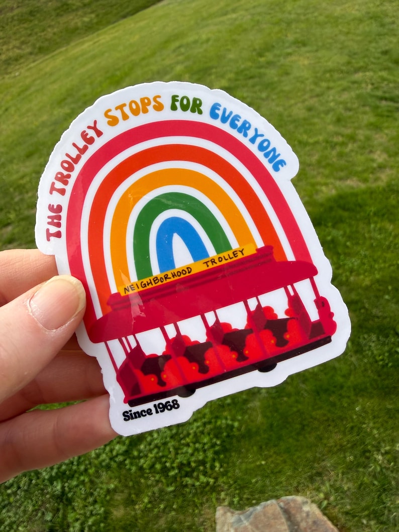 Pu&ograve; includere: Un adesivo colorato raffigurante un tram rosso con passeggeri, su uno sfondo arcobaleno. Il testo recita "THE TROLLEY STOPS FOR EVERYONE" e "NEIGHBORHOOD TROLLEY", con "Since 1968" in basso. L'adesivo &egrave; tenuto su uno sfondo erboso verde.