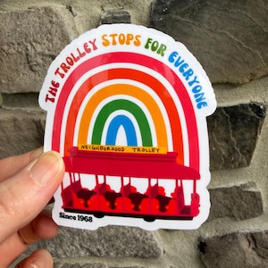 Pu&ograve; includere: Un adesivo colorato raffigurante un tram rosso sotto un arcobaleno. Il testo recita "The Trolley Stops For Everyone" e "Neighborhood Trolley". L'adesivo include anche "Since 1968".
