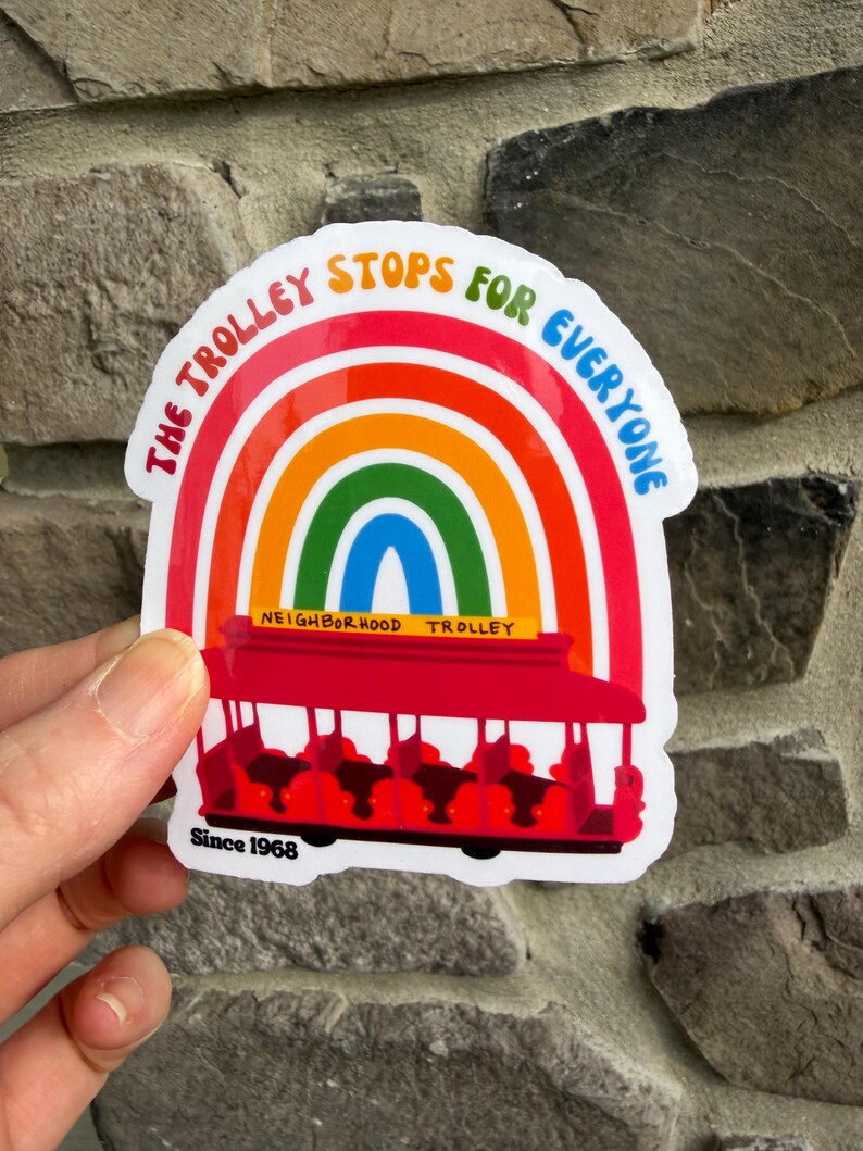 Pu&ograve; includere: Un adesivo colorato raffigurante un tram rosso sotto un arcobaleno. Il testo recita "The Trolley Stops For Everyone" e "Neighborhood Trolley". L'adesivo include anche "Since 1968".