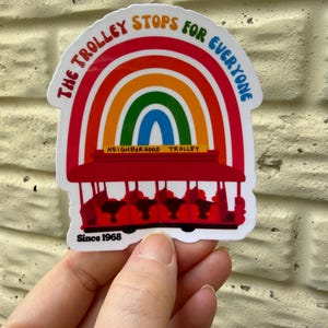 Pu&ograve; includere: Un adesivo colorato con un tram rosso con un arcobaleno sopra. Il testo recita "The Trolley Stops for Everyone" e "Neighborhood Trolley", con "Since 1968" sotto. L'adesivo &egrave; tenuto su uno sfondo strutturato.