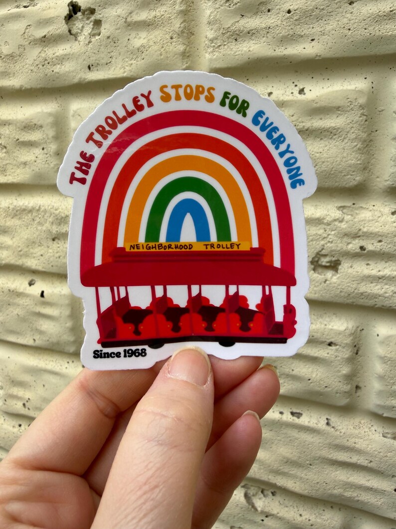 Pu&ograve; includere: Un adesivo colorato con un tram rosso con un arcobaleno sopra. Il testo recita "The Trolley Stops for Everyone" e "Neighborhood Trolley", con "Since 1968" sotto. L'adesivo &egrave; tenuto su uno sfondo strutturato.