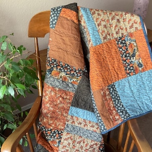 May include: A patchwork quilt featuring a variety of fabric patterns in shades of orange, brown, blue, and black. The quilt is constructed from rectangular and square panels with leaf motifs, geometric lines, and small circular patterns throughout.