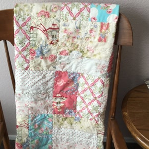 May include: A patchwork quilt featuring a variety of fabric squares with floral patterns, pink ribbons, and illustrations of figures in red hats. The quilt is constructed with visible stitching and displays a color palette of cream, pink, and light blue.