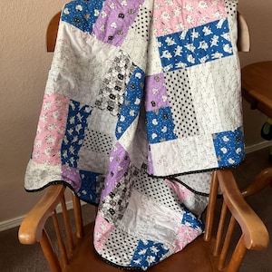 May include: A patchwork quilt featuring a repeating cat pattern in shades of pink, blue, purple, white, and black. The fabric squares are stitched together with a thin black border, showcasing various feline illustrations across the entire surface.