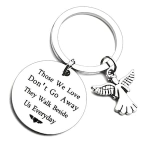 Op de afbeelding: Een zilverkleurige sleutelhanger met een ronde hanger en een engelbedel. De hanger heeft de tekst "Those We Love Don't Go Away They Walk Beside Us Everyday". De engelbedel heeft gedetailleerde vleugels en een vloeiende jurk.