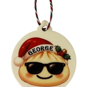 May include: A round, cream-coloured ornament featuring a cartoon dumpling wearing black sunglasses and a red Santa hat with the name GEORGE printed on the brim. The ornament includes a small sprig of holly and hangs from a red, green, and white cord.