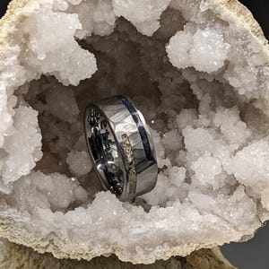 May include: A silver-toned ring with a textured surface and a dark blue inlay. The ring is set against a backdrop of white crystals within a geode, creating a striking contrast. The ring has the word "STRONGER" engraved on it.