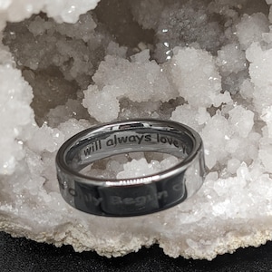 May include: A silver ring with the engraved words "will always love you" and "Only Begin". The ring is set against a backdrop of a white, crystalline structure.