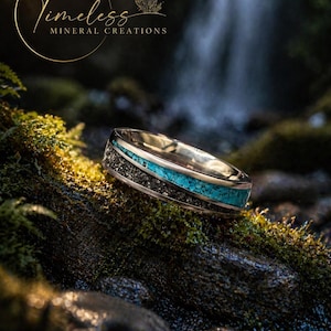 May include: A silver ring with a black stone inlay and a turquoise stone band. The ring is set against a mossy rock with a waterfall in the background. The ring is from Timeless Mineral Creations.